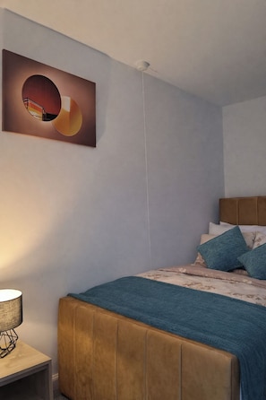 Un dormitorio con cama, cabecera, mesita de noche, lámpara y una obra de arte colgada en la pared.