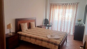 Habitación de hotel con cama, mesita de noche, lámpara, silla, escritorio, televisor y ventana con cortinas.