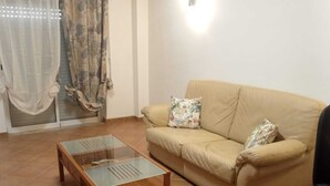 Una sala de estar con un sofá color beige, dos cojines estampados, una mesa de centro de vidrio y una silla de oficina negra.