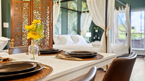 Ein Essbereich mit einem Tisch für zwei Personen, einer Vase mit gelben Blumen und einem Bett mit Baldachin im Hintergrund.