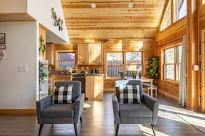 Ein Interieur einer Holzhütte mit Holzwänden, einer Küchenzeile, zwei grauen Sesseln und einem Esstisch.
