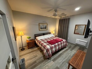 Un dormitorio con cama, lámparas de noche, ventilador de techo, ventana con cortinas, televisor de pared y radiador.
