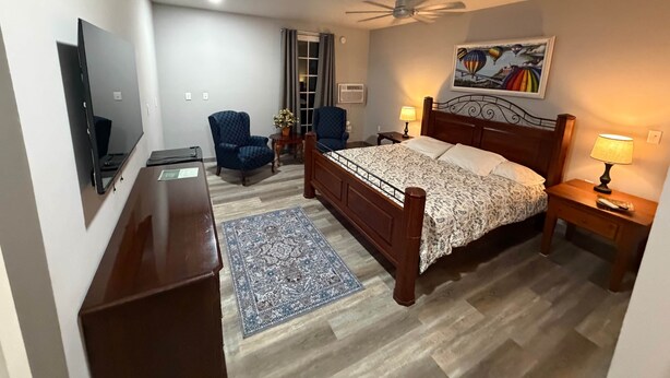 Un dormitorio con una cama, dos sillas, un escritorio, un televisor, un ventilador de techo y un cuadro en la pared.
