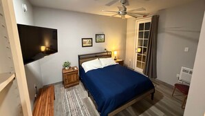 Quarto com cama, ventoinha no teto, TV fixada na parede, mesa de cabeceira com abajur e janela com cortinas.