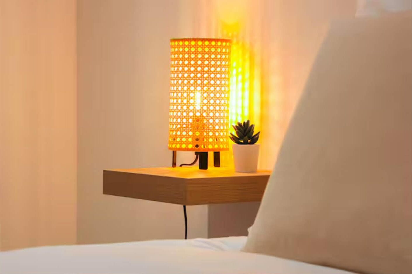 Une table de chevet avec une lampe, une petite plante en pot et un oreiller blanc.
