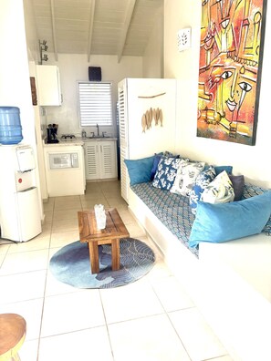 Een knus woongedeelte met een kitchenette, een bank met opvallende kussens, een houten salontafel en een groot abstract schilderij aan de muur.