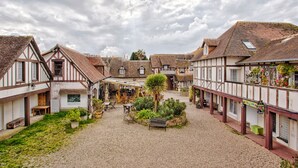A courtyard with traditional half-timbered buildings, a central seating area, and potted plants. - L'Escale Bleue - Cosy & Balneo, Village & Sea (Dives-sur-Mer)