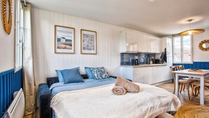A bedroom with a bed, a small table, and a kitchenette in the background. - L'Escale Bleue - Cosy & Balneo, Village & Sea (Dives-sur-Mer)