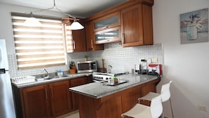 A kitchen with wooden cabinets, a granite countertop, a microwave, a stove, and a sink. There are two white chairs and a hanging light fixture. - Spacious 3-bedroom apartment in serene Santiago de los Caballeros with AC (Santiago de los Caballeros)