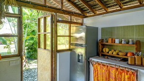 A compact kitchen with a refrigerator, a window, and a shelf with kitchenware. - Charming 1BR Garden Bungalow (Temae)