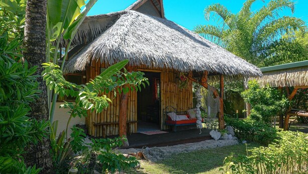 A thatched-roof hut with a bed and a small porch, surrounded by tropical vegetation. - Charming 1BR Garden Bungalow (Temae)