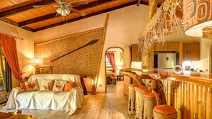 A rustic interior with a wooden ceiling, a bed with white bedding, a woven wall, and a kitchen area.
