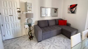 A living room with a grey sectional sofa, a glass coffee table, a white door, and a marble floor. - 1 BR Sea View · Steps to Beach · Los Cristianos (Los Cristianos)