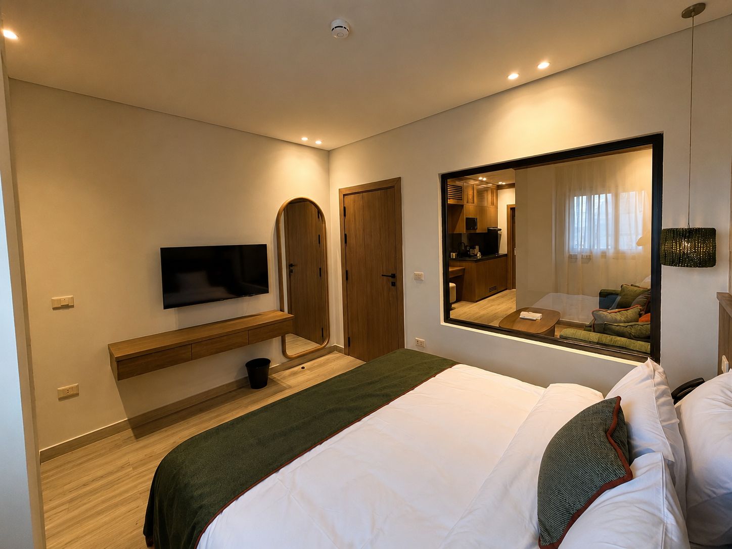 A modern hotel room with a bed, a TV, a mirror, and a view of the kitchen.