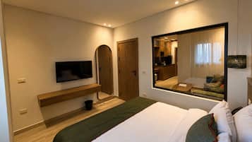 A modern hotel room with a bed, a TV, a mirror, and a view of the kitchen.