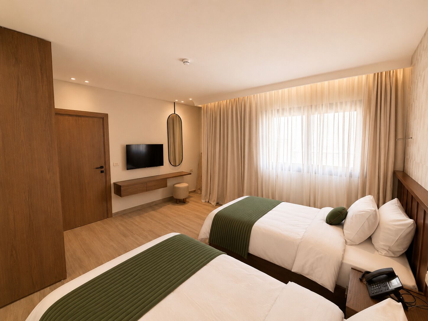 A hotel room with two beds, a TV, a bedside table, a lamp, and a window with curtains.