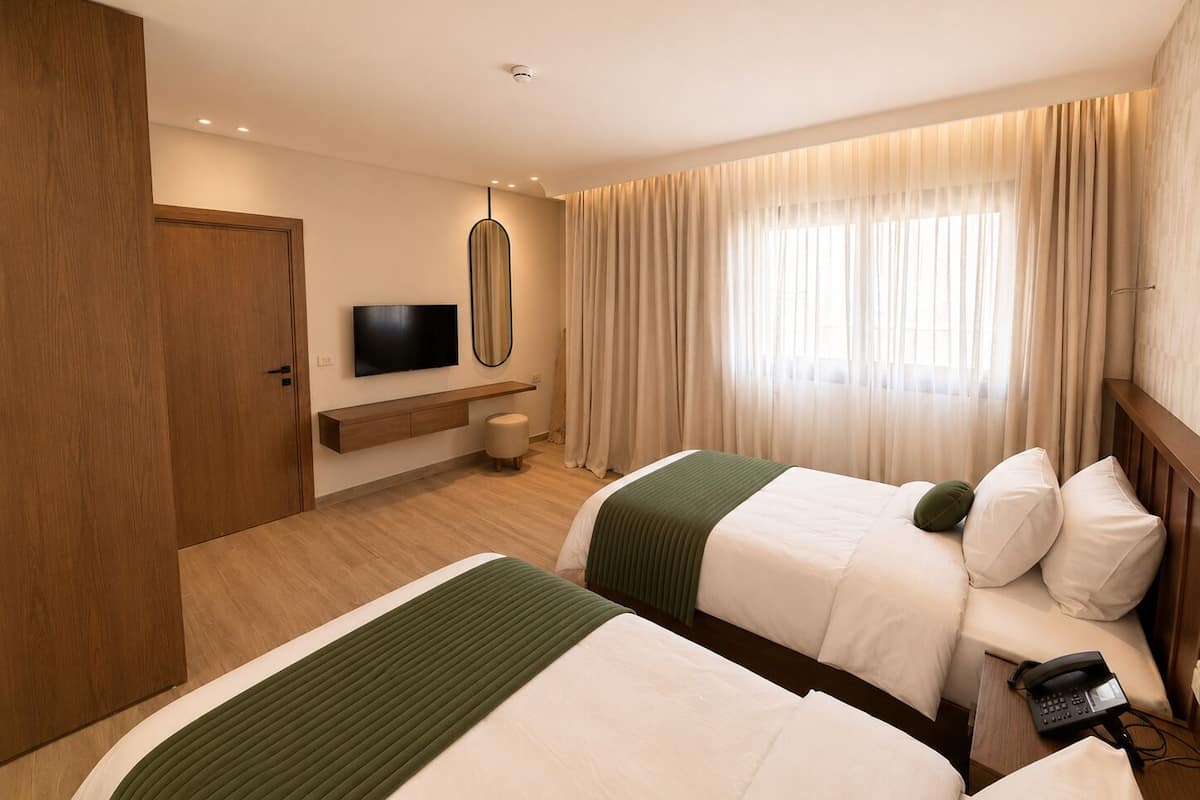 Una habitación de hotel con dos camas, televisor, mesita de noche, lámpara y ventana con cortinas.