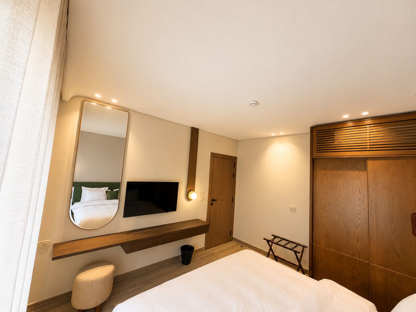 A hotel room with a bed, a television mounted on the wall, a wooden wardrobe, and a small stool.
