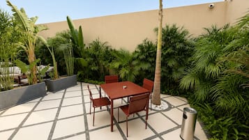 An outdoor patio with a table and chairs, surrounded by lush greenery and a tiled floor.