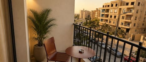 A balcony with two chairs and a table, a potted plant, and a view of a cityscape with buildings and a street.