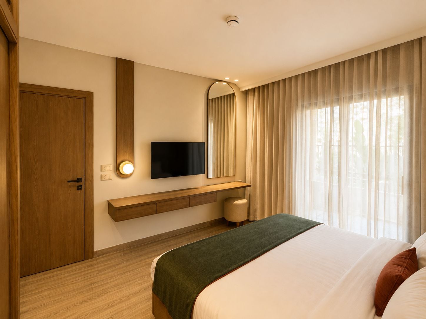 A hotel room with a bed, a television mounted on the wall, a wooden door, and a window with curtains.