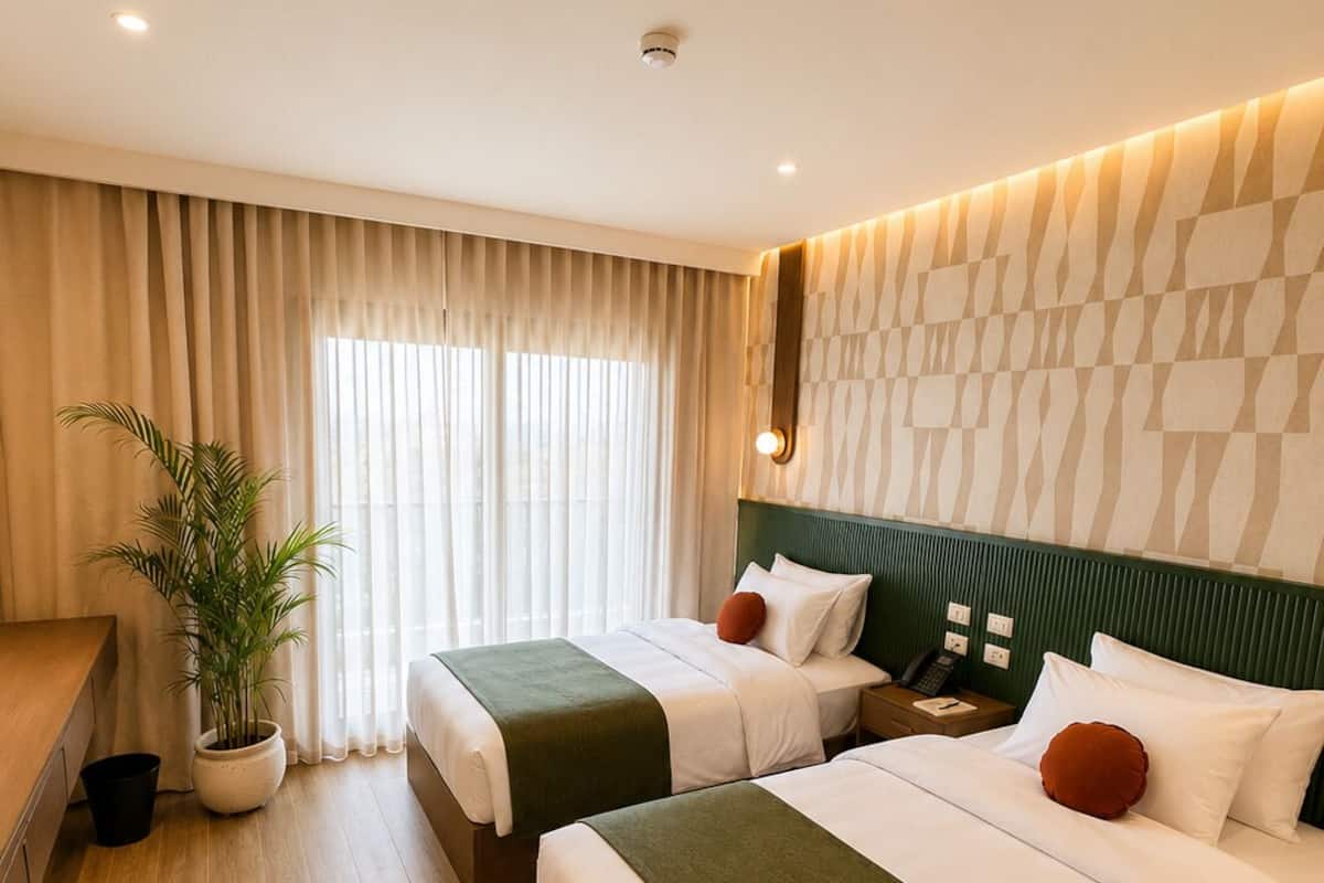 Habitación de hotel con dos camas, un escritorio, una silla, una planta en maceta y un ventanal con cortinas.