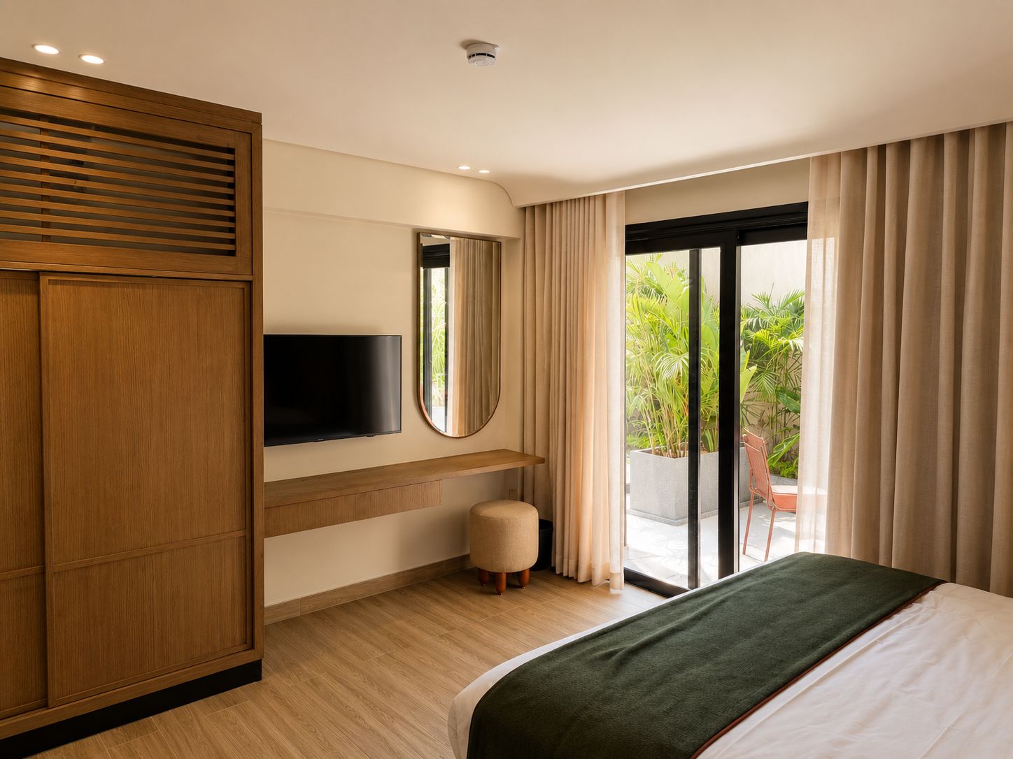 Een hotelkamer met een bed, een televisie, een spiegel, een klein krukje en een schuifglazen deur die leidt naar een balkon met planten.