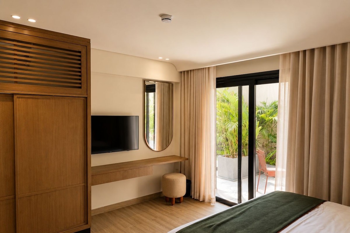 A hotel room with a bed, a television, a mirror, a small stool, and a sliding glass door leading to a balcony with plants.