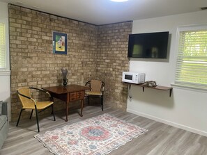 A dining area with a brick wall, a wooden table, two chairs, a decorative rug, a mounted TV, and a microwave on a shelf. - Spacious and marvelous studio with AC in quiet urban setting (Raleigh)