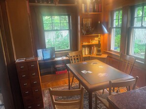 A room with a desk, a bookshelf, a window with a view of greenery, and a lamp.