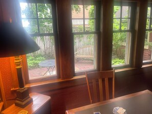 A room with a table, a chair, a lamp, and a window with a view of a garden and a wooden bench.