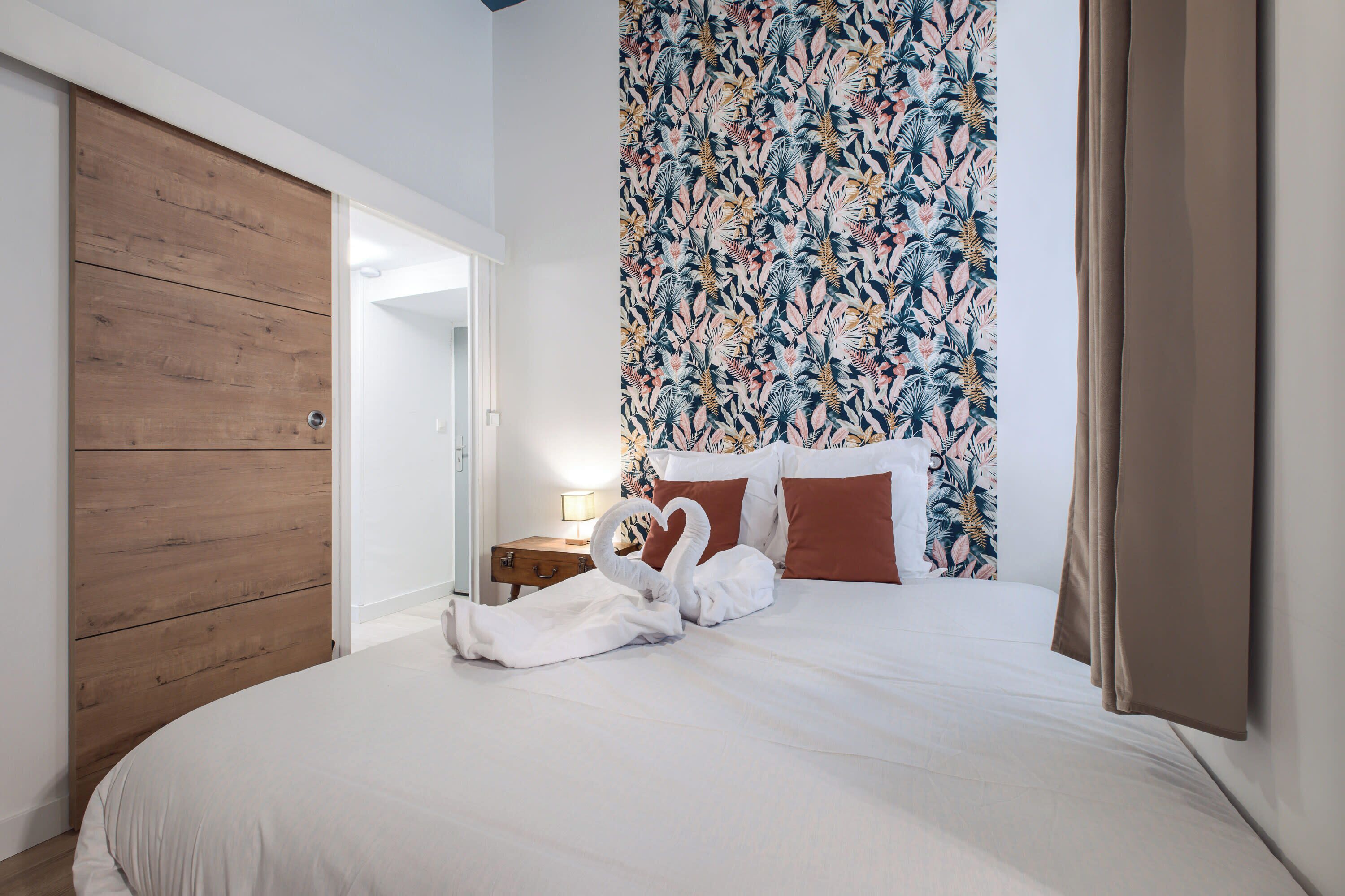 Quarto com guarda-roupa de madeira, parede com padrão floral, cama com roupa de cama branca e mesa de cabeceira com abajur.