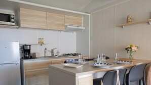 A modern kitchen with wooden cabinets, a built-in microwave, a coffee maker, and a dining area with chairs and a table set for four. - Oléon - Confort 2 TV – 5p  (2) (Saint-Georges-d'Oléron)