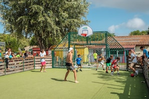 An outdoor basketball court with a green fence, spectators, and a clear sky.