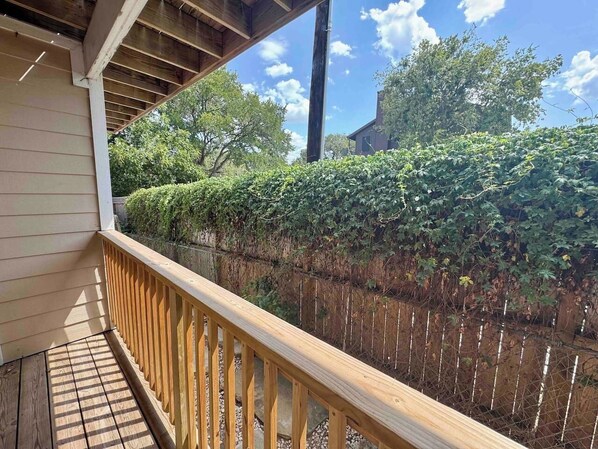 A wooden deck with a metal railing, surrounded by a hedge and a wooden fence. - Wasserlodge #104 | Sleeps 4 | Comal Riverfront (New Braunfels)