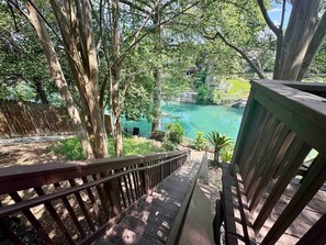 A wooden deck with a railing overlooking a turquoise pool surrounded by trees. - Wasserlodge #104 | Sleeps 4 | Comal Riverfront (New Braunfels)