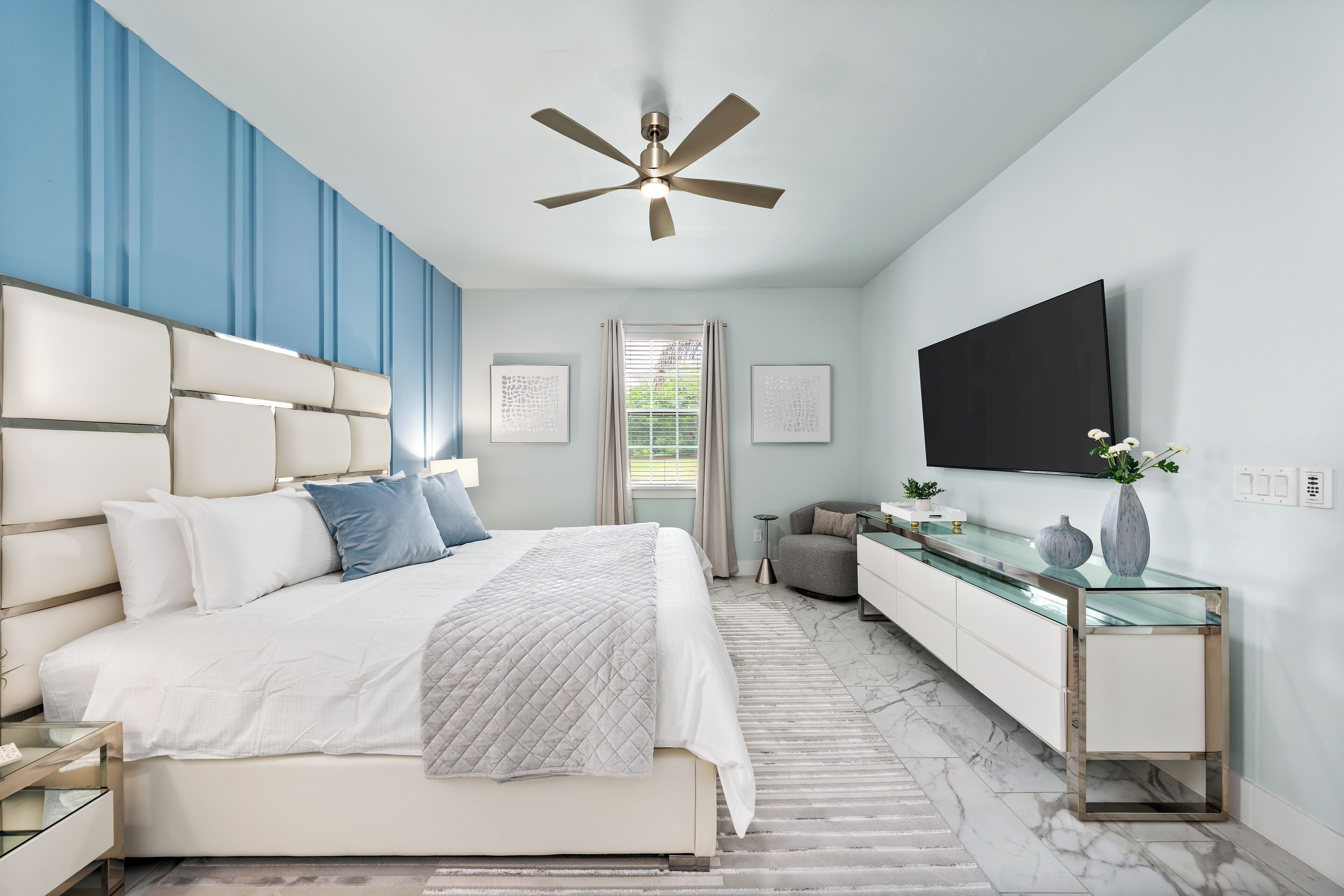 A modern bedroom with a bed, bedside tables, a glass console table, a ceiling fan, and a TV mounted on the wall.