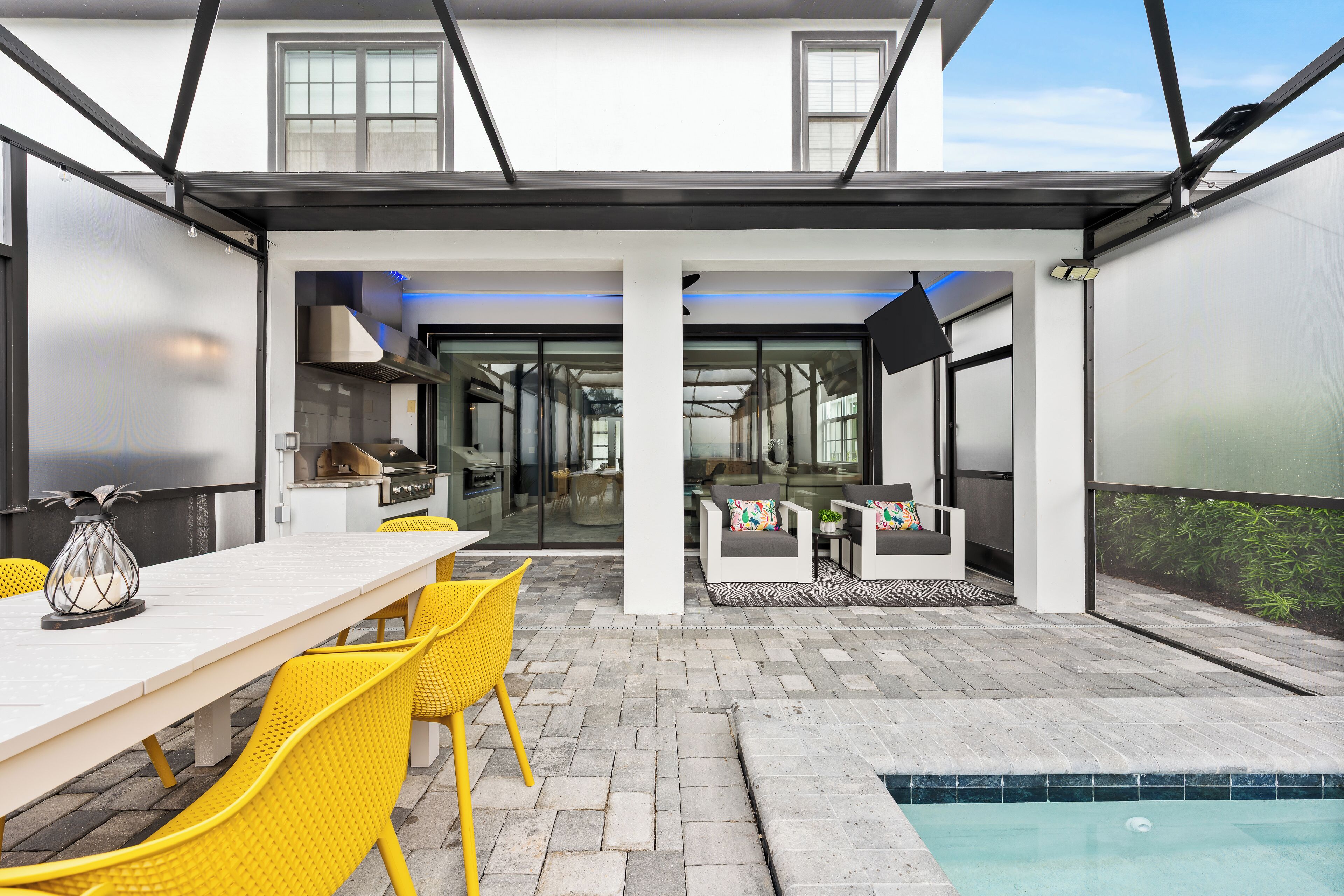 A modern outdoor patio with a dining area, yellow chairs, a pool, and a view into the house.