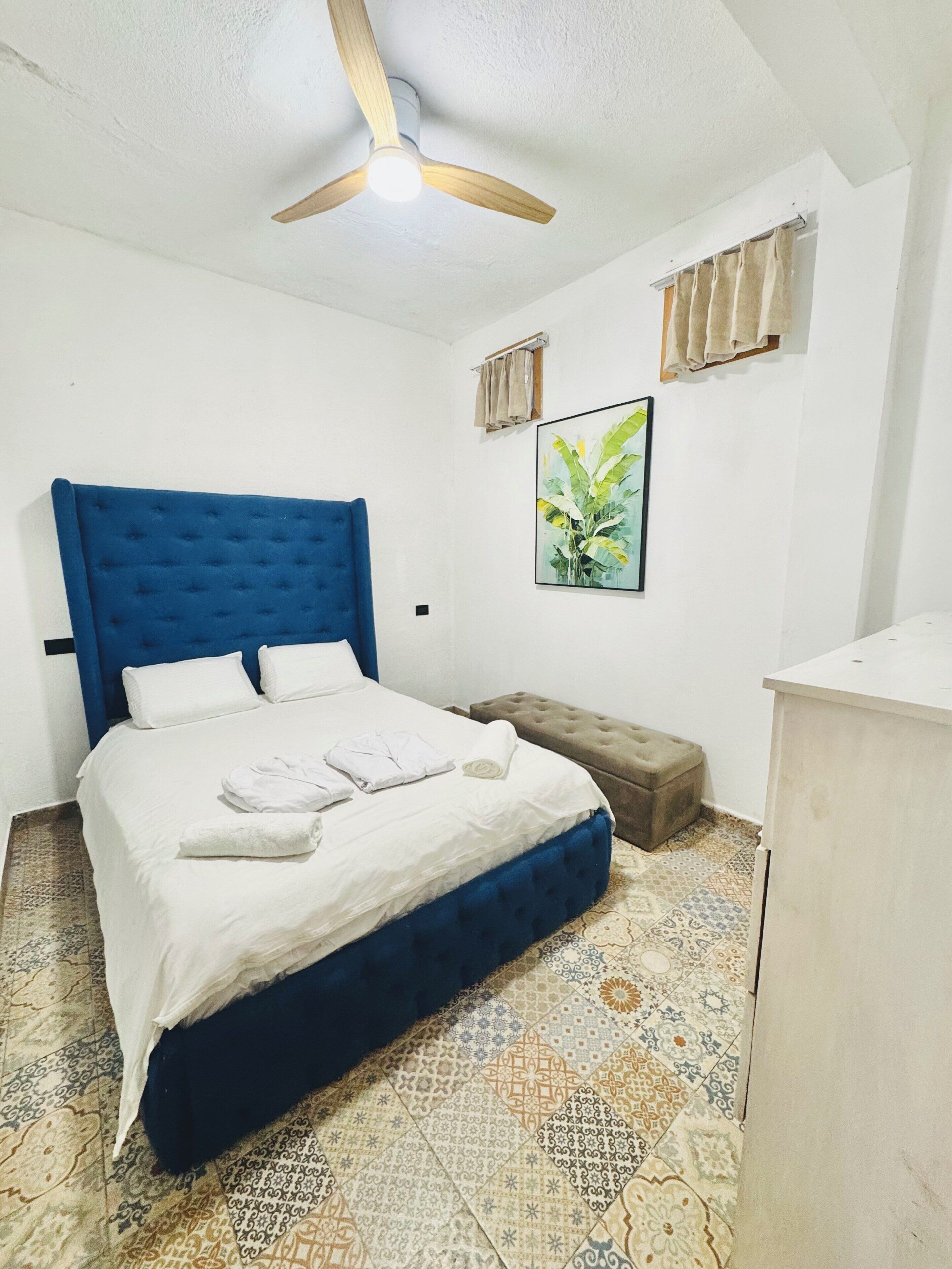 Un dormitorio con cabecera azul, cama con ropa blanca, reposapiés color beige, ventilador de techo y un cuadro en la pared.