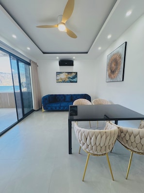 A modern room with a ceiling fan, a dining table with chairs, a blue sofa, and a large window with a view.
