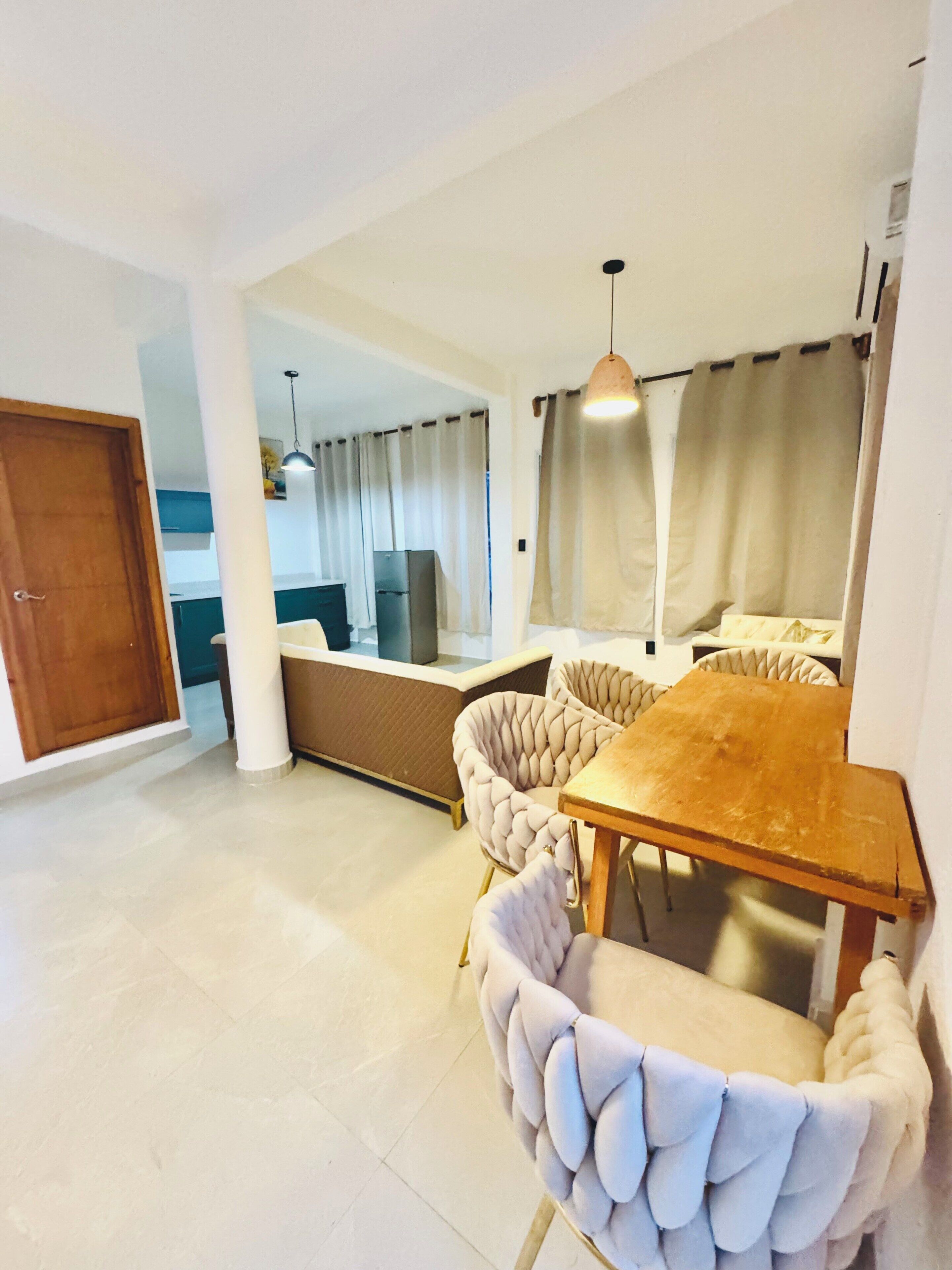 Comedor con mesa de madera y sillas acolchadas, cocina al fondo y una puerta a la izquierda.