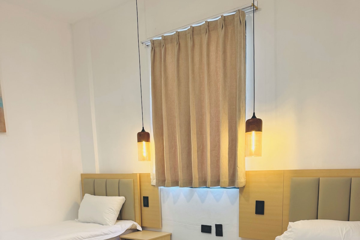 Een hotelkamer met een groot bed, een kleiner bed, een nachtkastje en een raam met gordijnen.