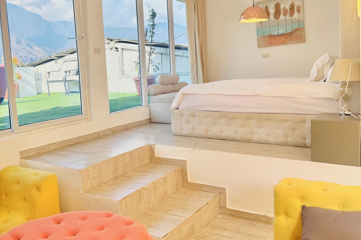 Een slaapkamer met een bed, een bank, een stoel en uitzicht op een berglandschap door grote ramen.