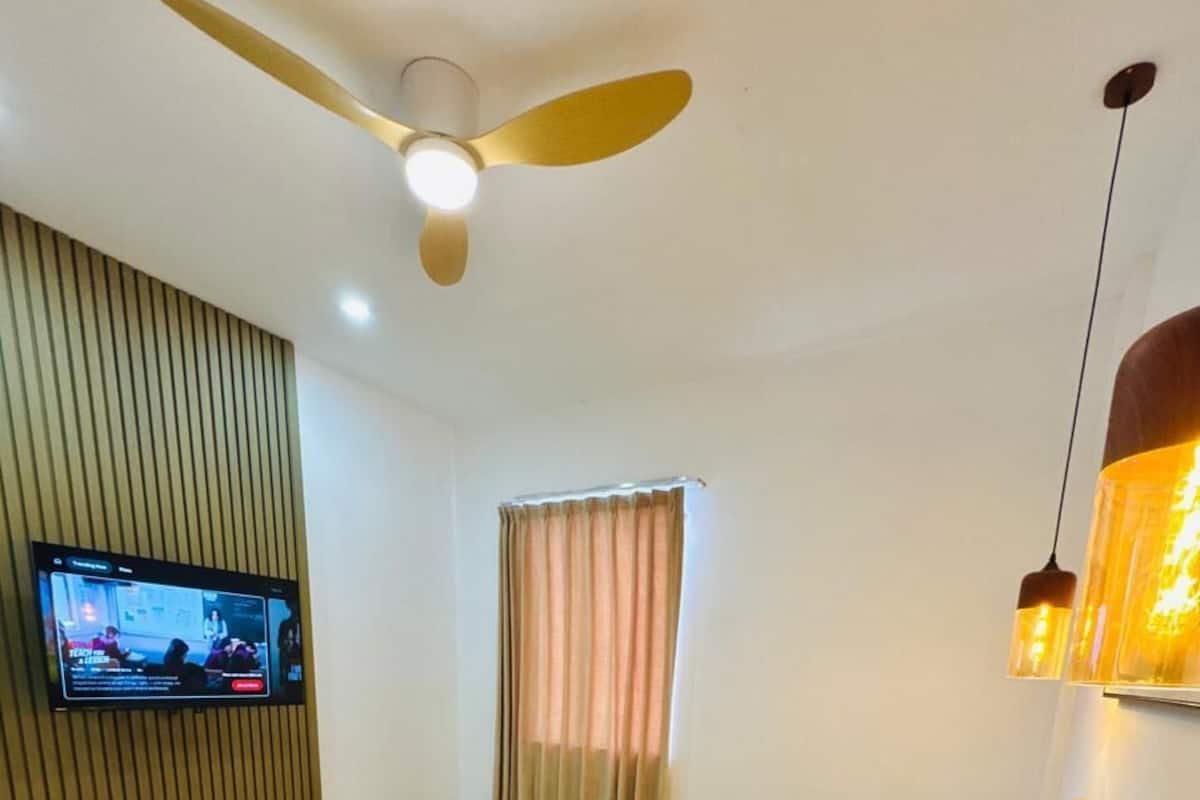 Egy szállodai szoba, amelyben található egy ágy, egy falra szerelt televízió, egy mennyezeti ventilátor és egy függönnyel ellátott ablak.