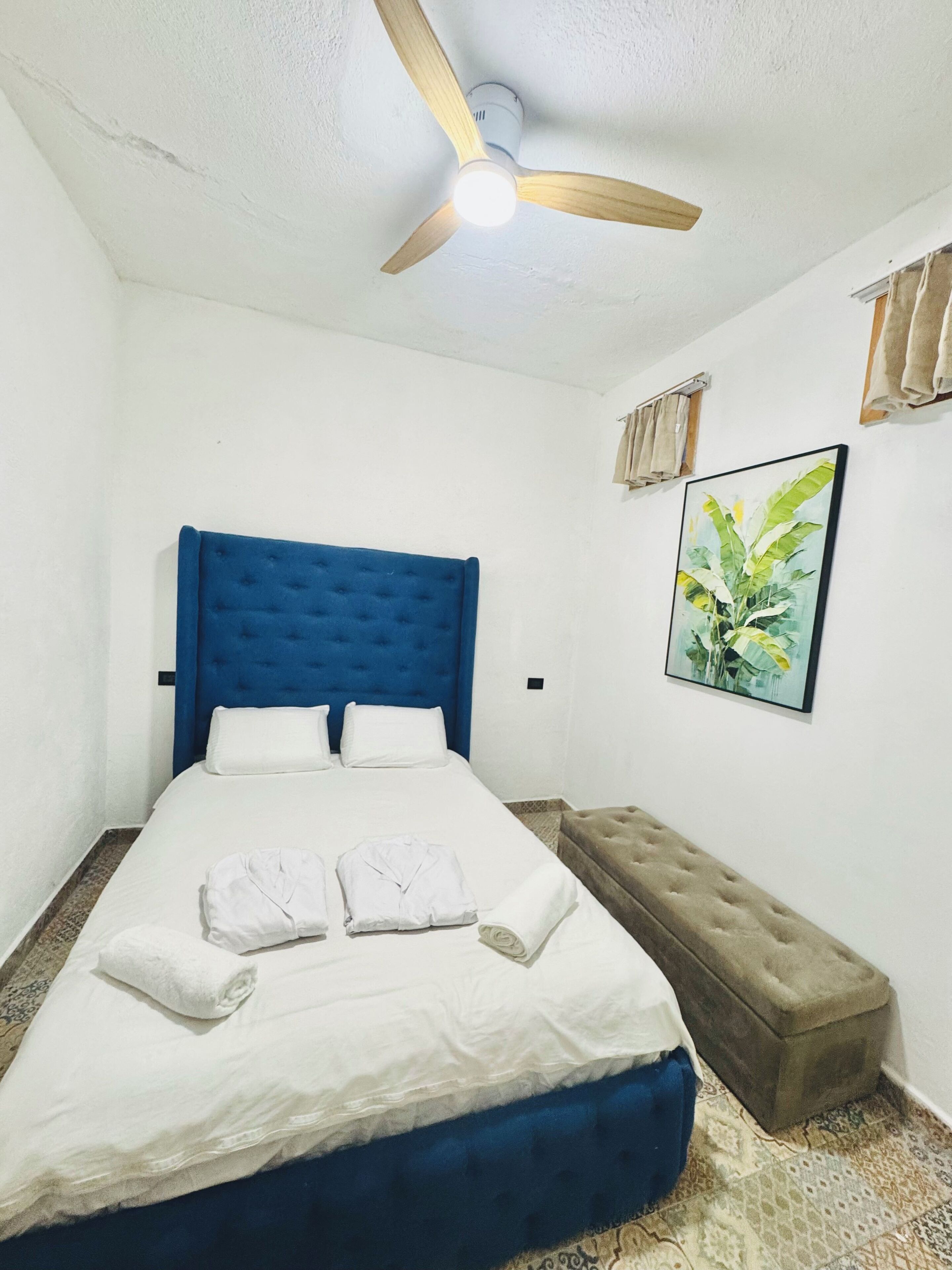 Un dormitorio con cama, ventilador de techo, banco, pintura y ventana con cortinas.