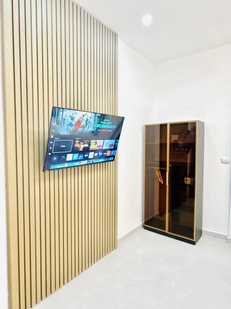 Quarto com televisão fixada na parede, guarda-roupa com porta de vidro e chão em azulejos.