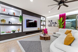 Ein modernes Wohnzimmer mit Flachbildfernseher, Deckenventilator, einem weißen Sofa und einem Bücherregal mit Dekoration.