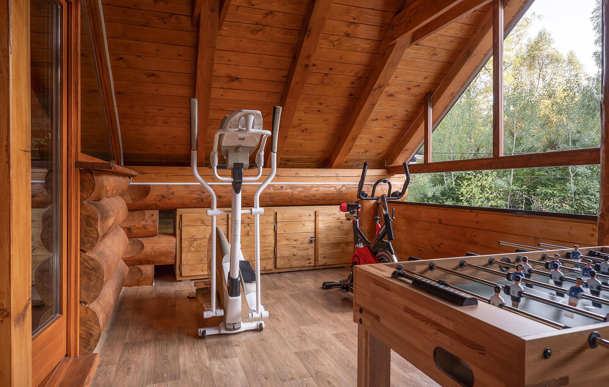 A wooden cabin interior with a stationary bike, a foosball table, and a large window.