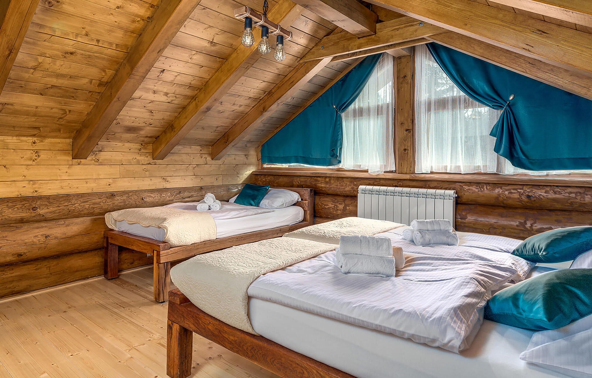 A cozy attic bedroom with wooden beams, two beds, a window with curtains, and a hanging light fixture.