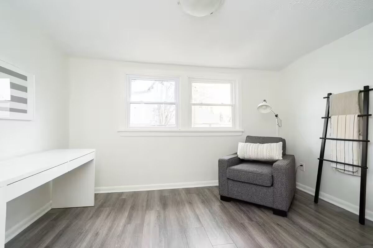 A room with a white desk, two windows, a gray sofa, a lamp, and a black rack.